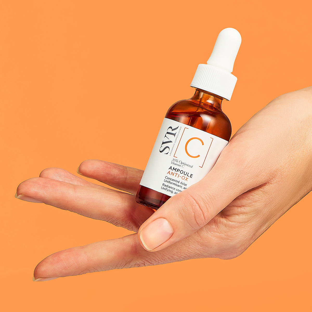 Mano che tiene un flacone in vetro ambrato di SVR Ampoule Anti-Ox con contagocce bianco su sfondo arancione.
