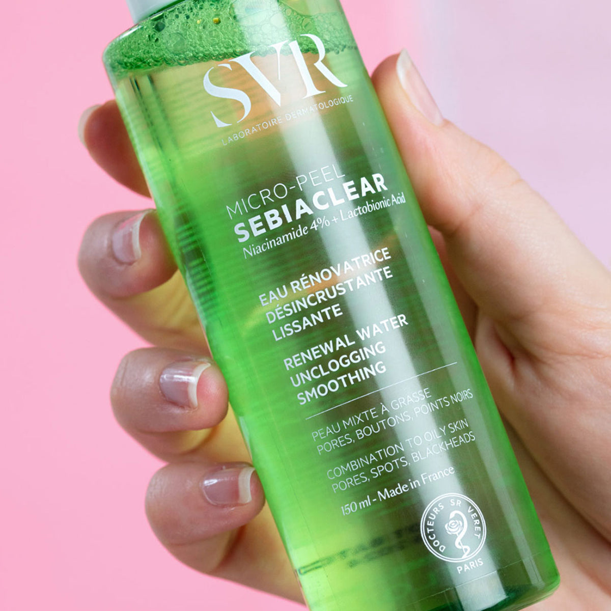 Flacone trasparente da 150ml di SVR Sebiaclear Micro-Peel con liquido verde, tenuto in mano su sfondo rosa.