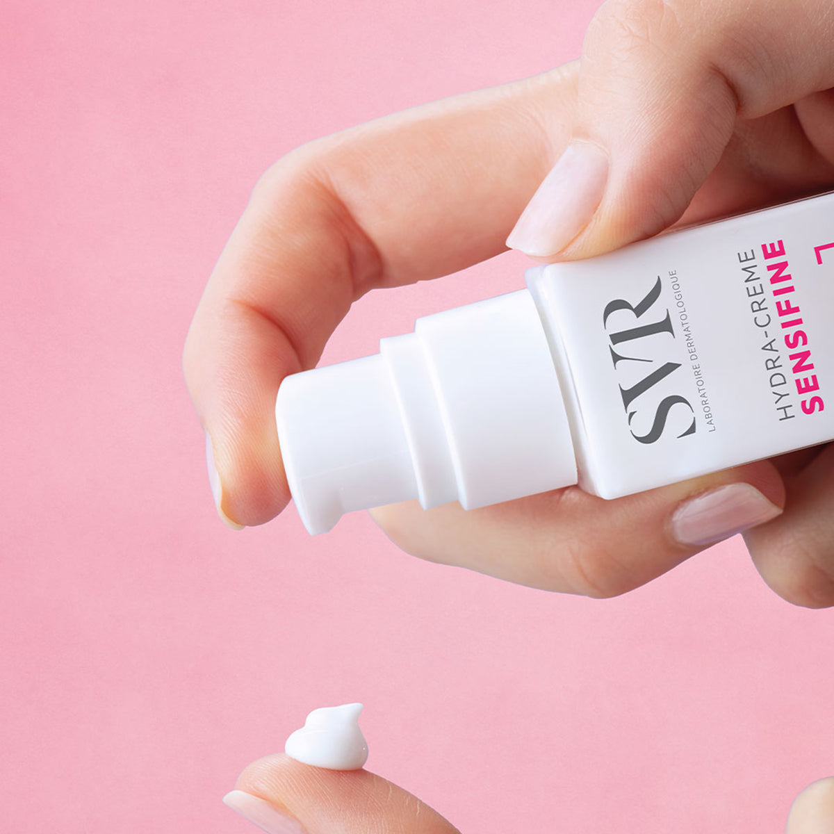 Mano che eroga la crema idratante bianca SVR Sensifine Hydra-Crème da un flacone con dosatore su sfondo rosa.