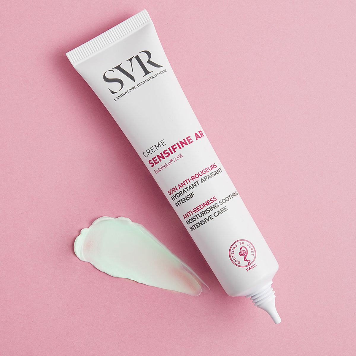 Tubo bianco di crema SVR Sensifine AR con una goccia di gel verde su sfondo rosa, ideale per pelli soggette ad arrossamenti.