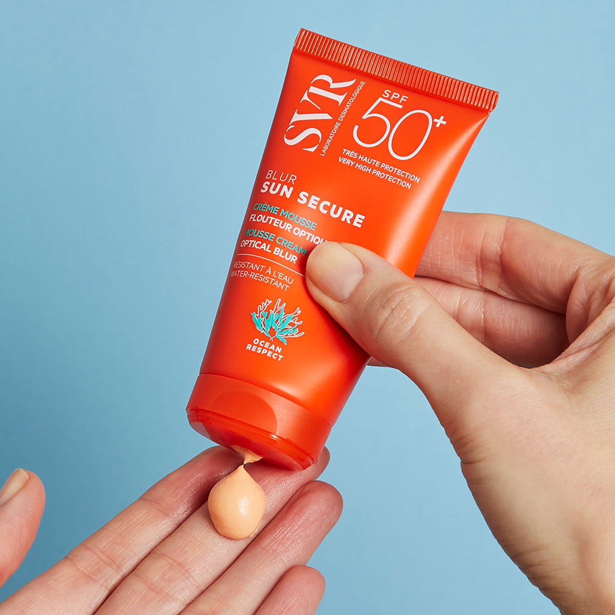 Tubo arancione di SVR Sun Secure Blur SPF50+ che eroga una crema mousse color albicocca sul palmo di una mano.
