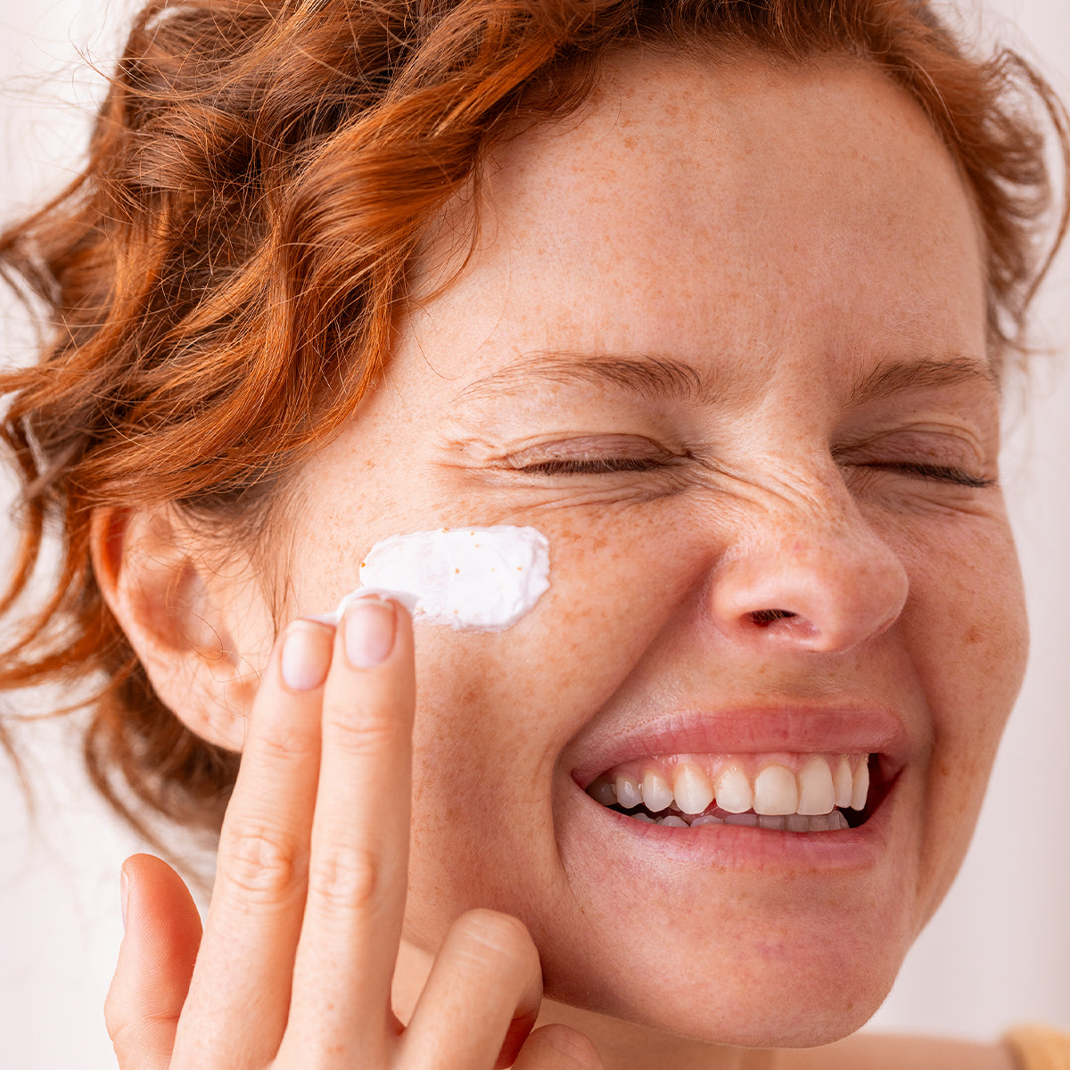 Donna dai capelli rossi che applica una crema viso bianca e cremosa sulla guancia, sorridendo con un'espressione radiosa.