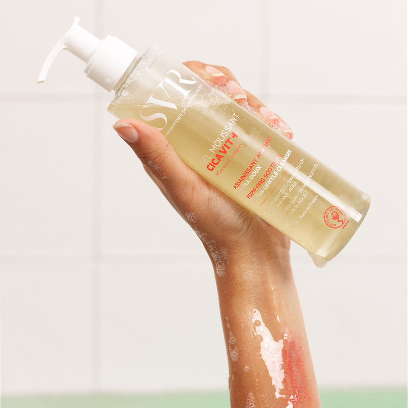 Flacone trasparente con dosatore di SVR Cicavit+ Gel Moussant tenuto in mano su sfondo piastrellato bianco.