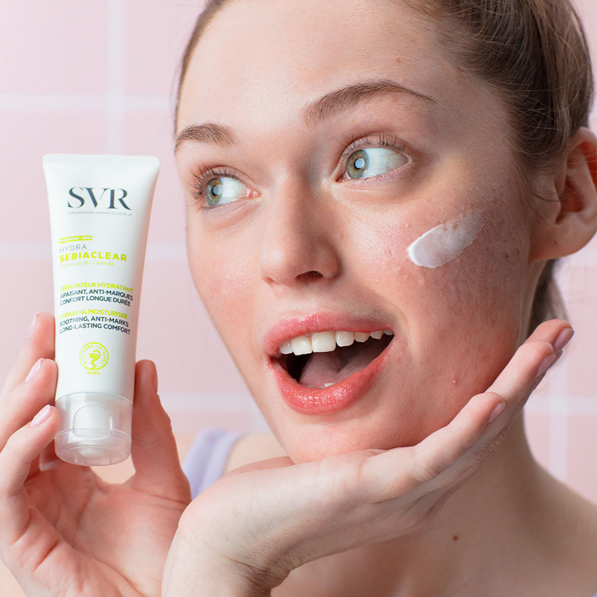 Donna che tiene in mano il tubetto bianco di SVR Sebiaclear Hydra con crema idratante applicata sulla guancia.