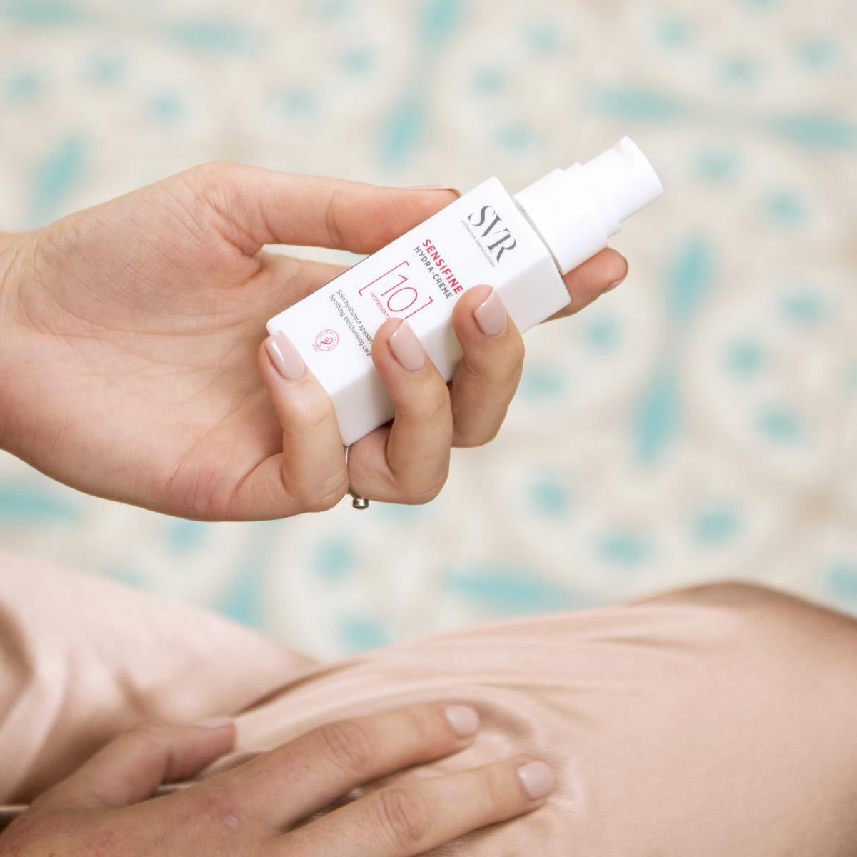 Mano che tiene il flacone bianco di SVR Sensifine Hydra-Crème con erogatore a pompa su sfondo sfocato.