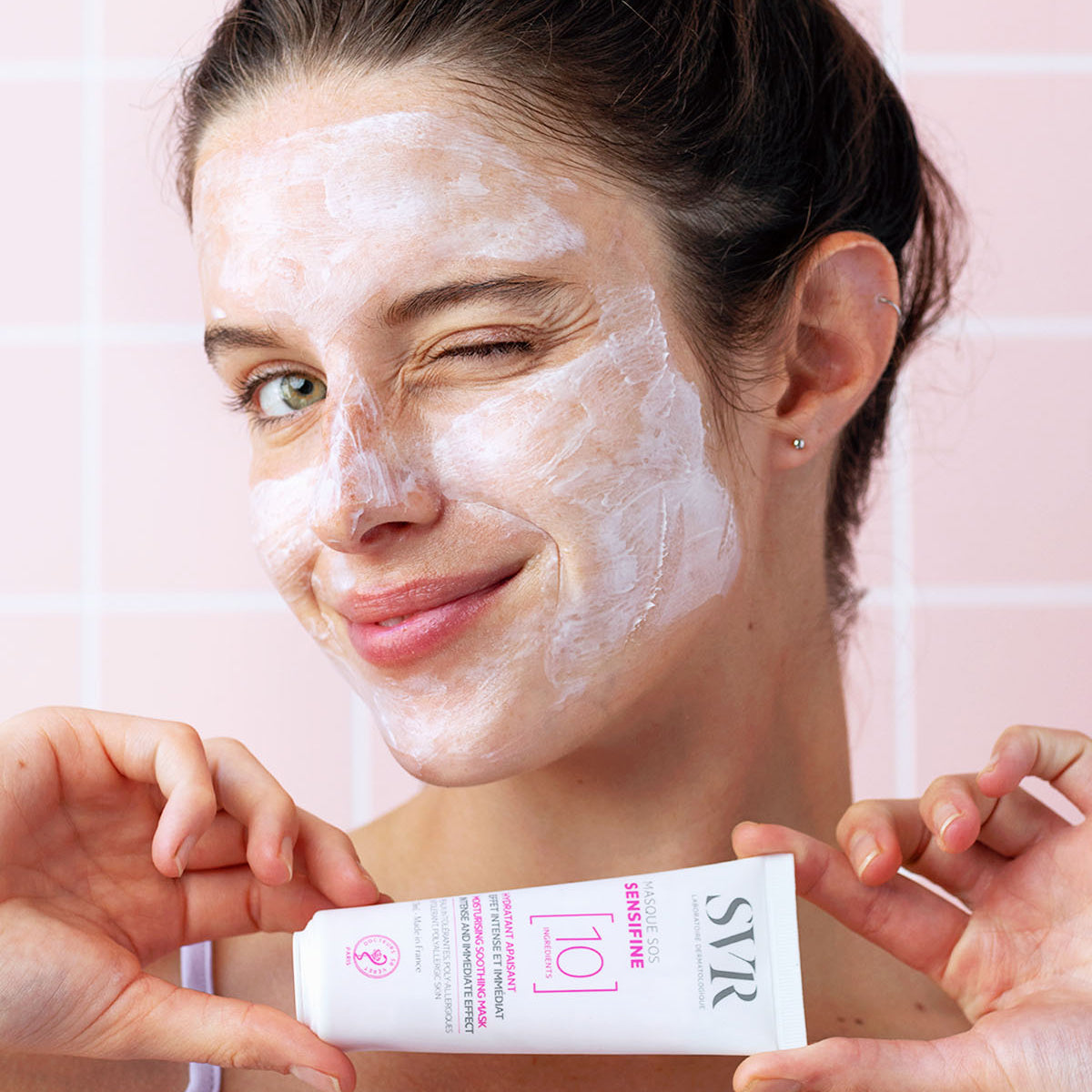 Donna che applica la maschera viso lenitiva SVR Sensifine SOS su pelle irritata, tenendo in mano il tubetto bianco del prodotto.