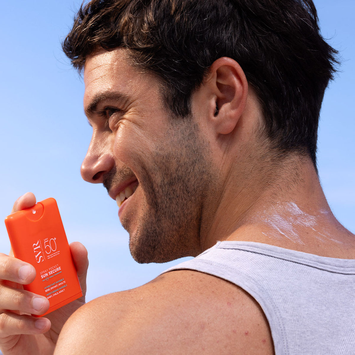 Uomo sorridente tiene in mano un flacone spray tascabile arancione SVR Sun Secure SPF50+ contro un cielo azzurro.