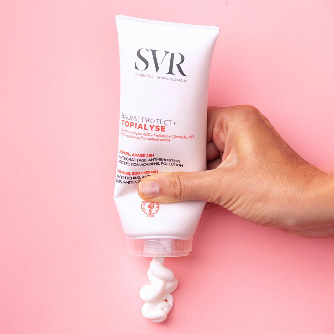 Mano che preme un tubo bianco di balsamo SVR Topialyse Protect+ su sfondo rosa, mostrando la texture cremosa del prodotto.