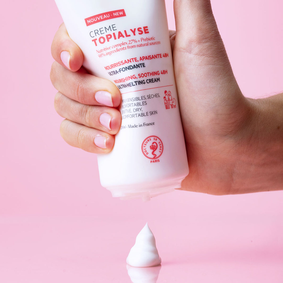 Mano che preme un tubo bianco di crema Topialyse su sfondo rosa, con una goccia di prodotto vellutato che cade verso il basso.