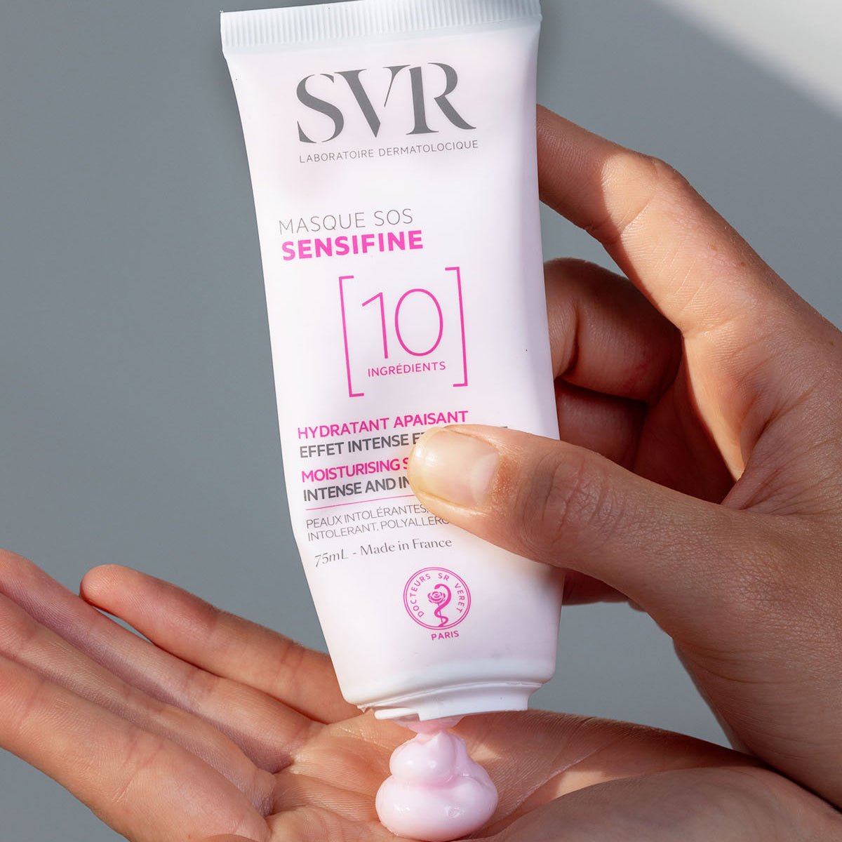 Tubo bianco di SVR Sensifine Masque SOS da 75ml che eroga una crema lenitiva di colore rosa tenue sul palmo della mano.