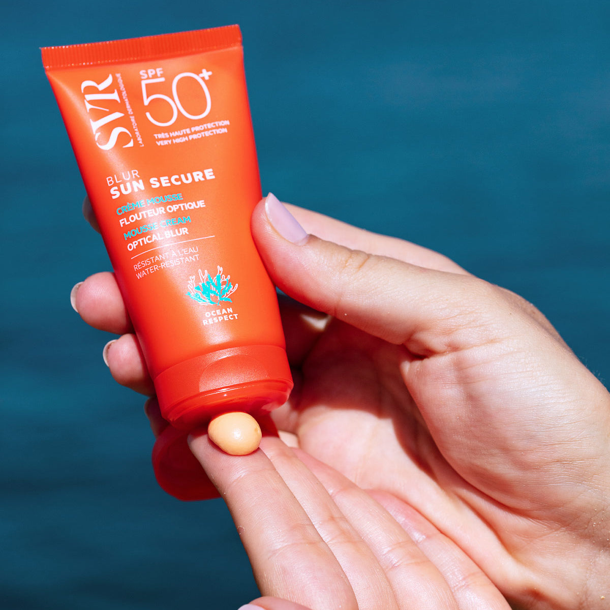 Tubo arancione di SVR Sun Secure Blur SPF50+ con texture in mousse color albicocca applicata sulla mano.