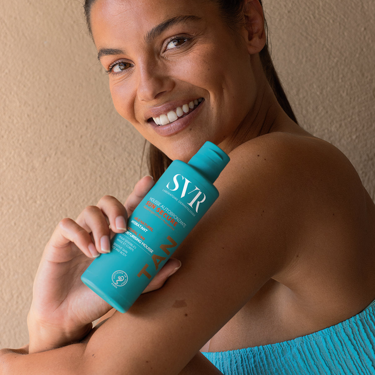 Donna sorridente che tiene in mano il flacone turchese di SVR Sun Secure Mousse Autobronzante per viso e corpo.