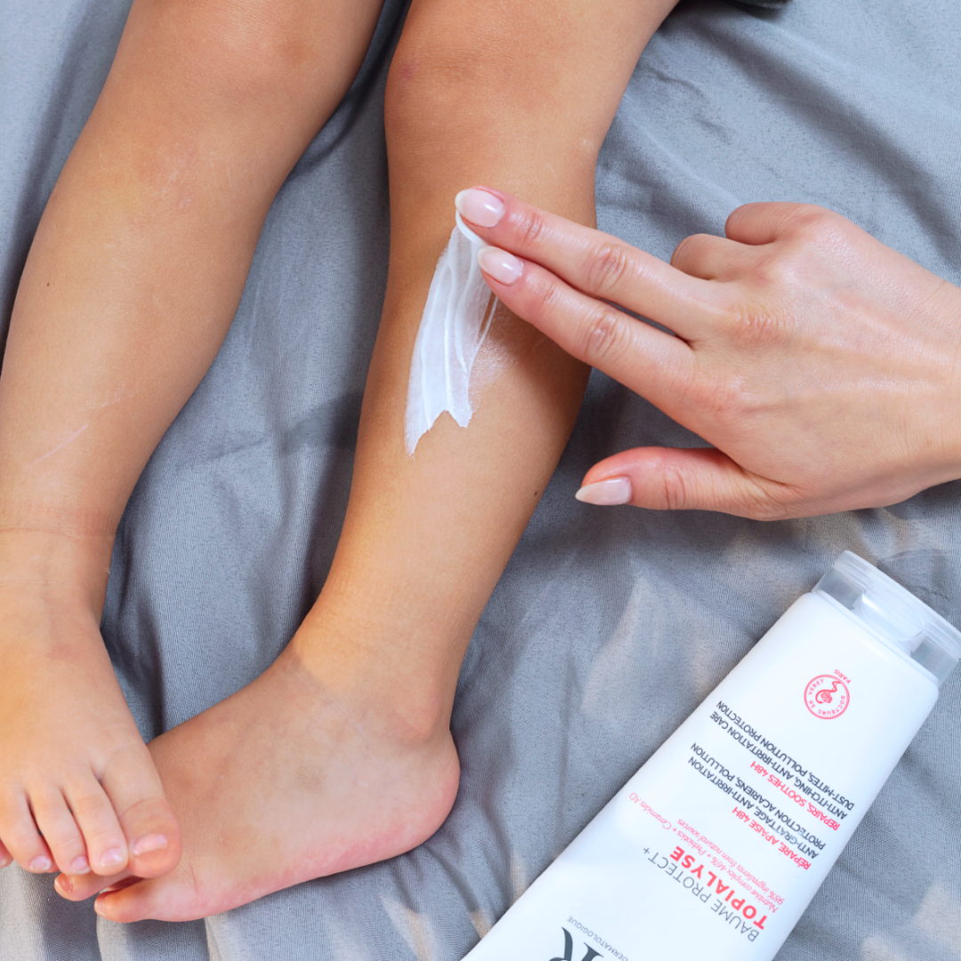 Mano che applica il balsamo bianco TOPIALYSE Baume Protect+ sulla gamba di un bambino su lenzuola grigie.