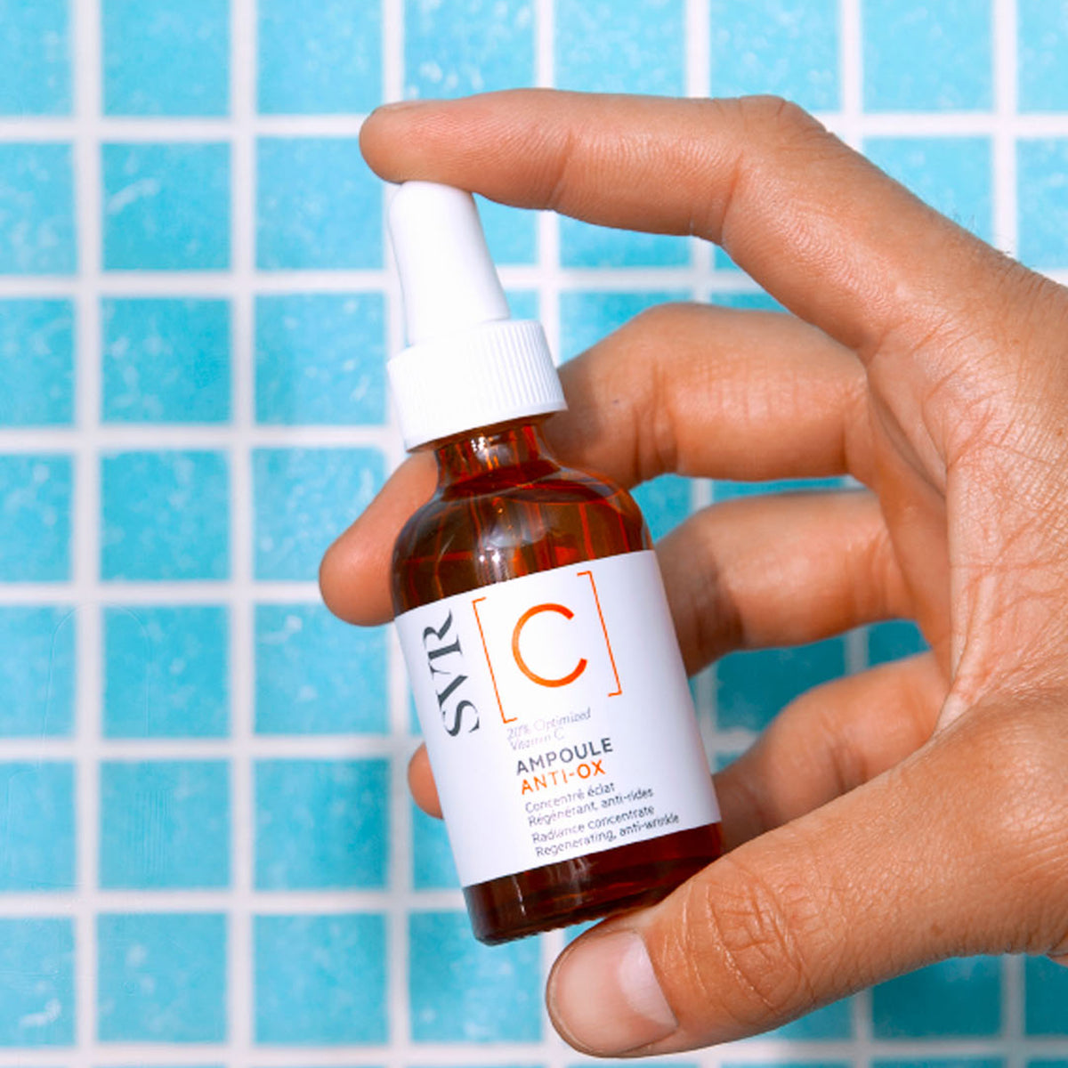 Flacone in vetro ambrato di SVR Ampoule Anti-Ox con contagocce bianco, tenuto in mano su sfondo di piastrelle azzurre.