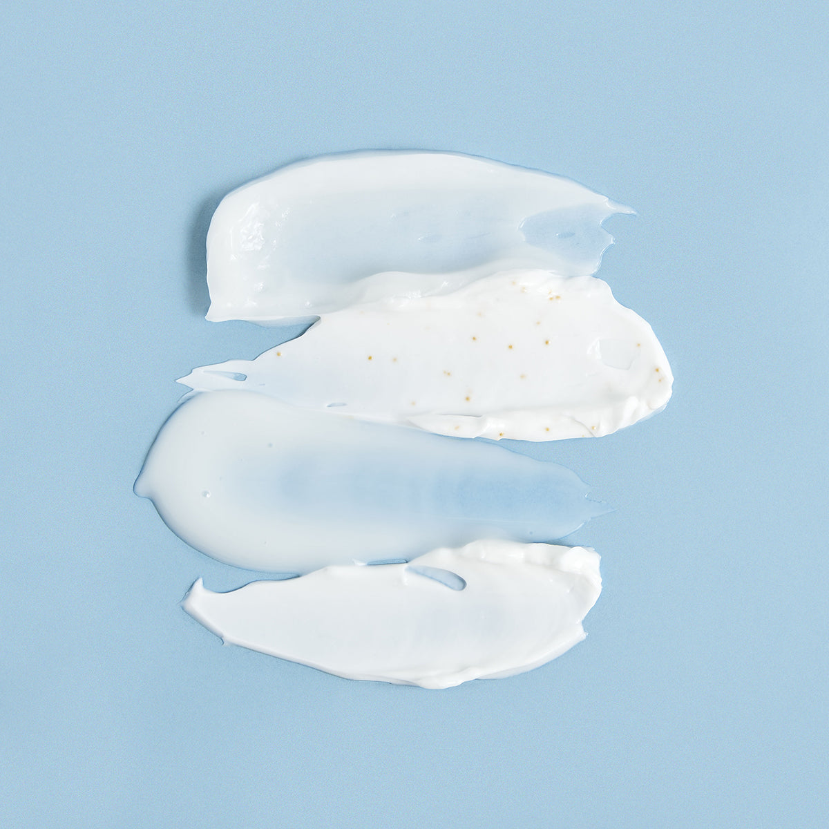 Tre strisce di crema viso bianca e gel trasparente su sfondo azzurro, con microgranuli dorati visibili nella texture.