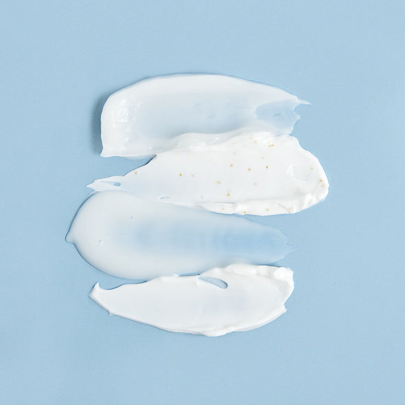 Tre strisce di crema viso bianca e gel trasparente su sfondo azzurro, con microgranuli dorati visibili nella texture.