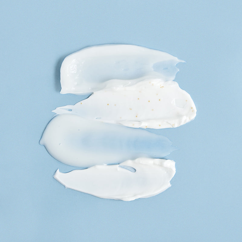 Tre strisce di crema viso bianca e gel trasparente su sfondo azzurro, con microgranuli dorati visibili nella texture.