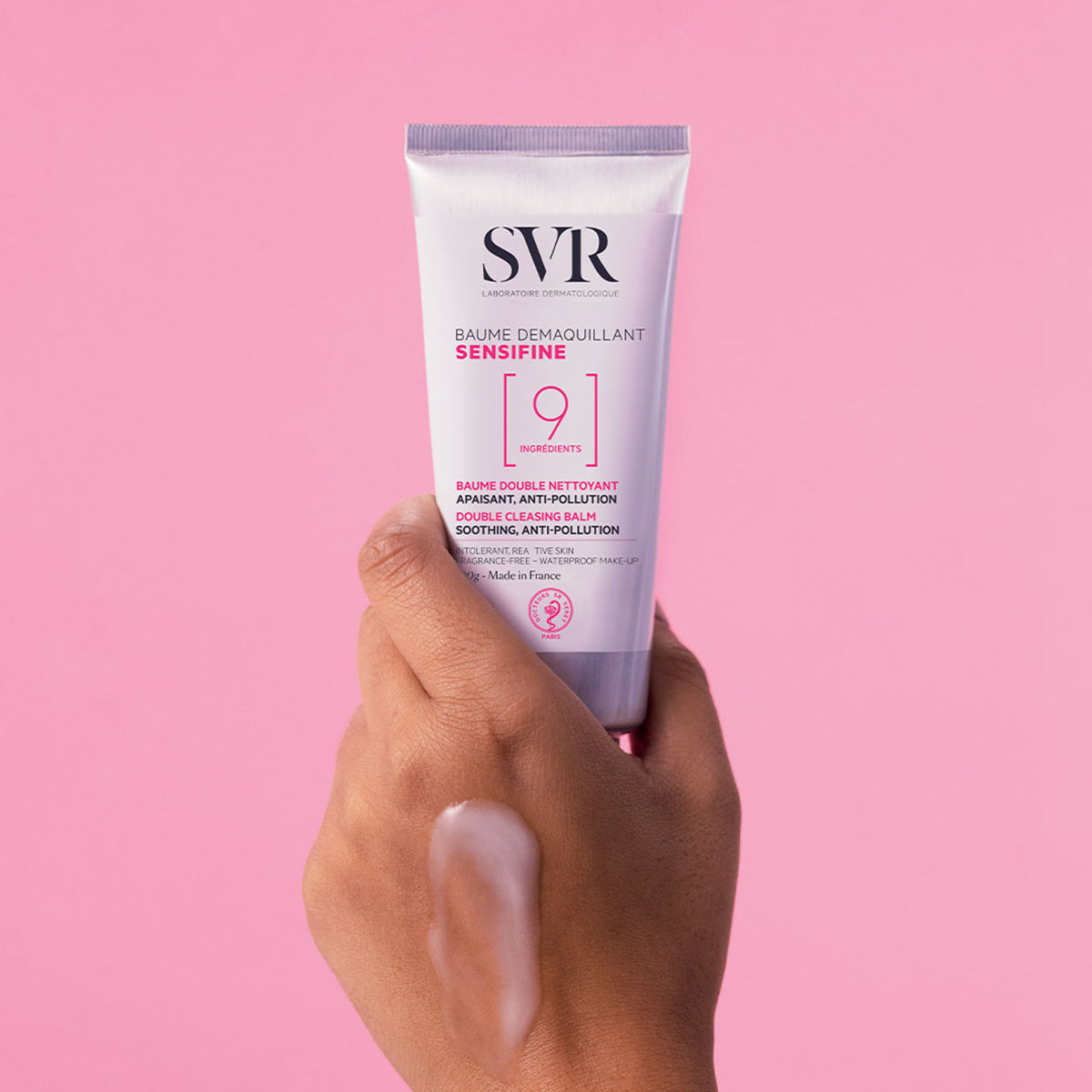 Mano che tiene il tubo bianco di SVR Sensifine Baume Démaquillant con texture balsamo-in-olio su sfondo rosa.