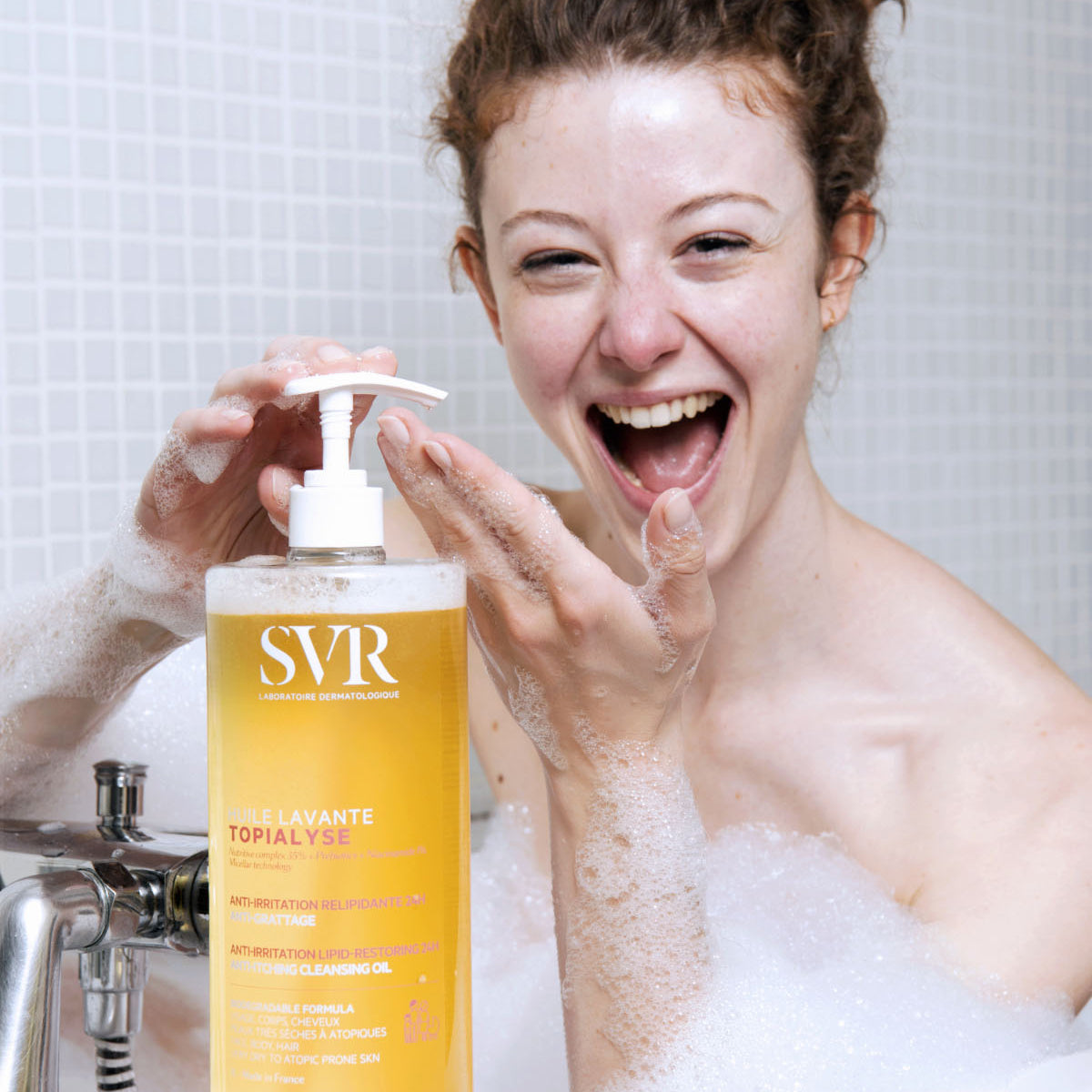 Donna sorridente nella vasca da bagno con flacone a pompa di SVR Topialyse Huile Lavante, olio detergente corpo schiumoso.