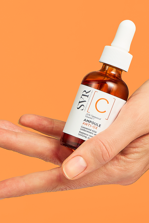 Mano che tiene un flacone in vetro ambrato di SVR Ampoule Anti-Ox con contagocce bianco su sfondo arancione.