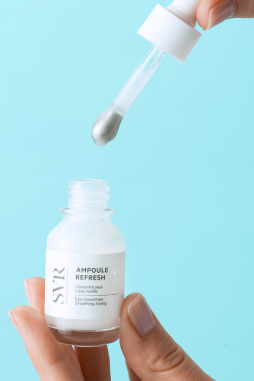 Siero contorno occhi SVR Ampoule Refresh in flacone di vetro con applicatore a pipetta in metallo su sfondo azzurro.
