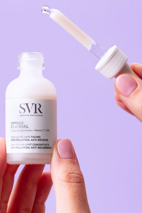Flacone di SVR Clairial Ampoule anti-macchie in vetro satinato tenuto in mano con contagocce su sfondo lilla.