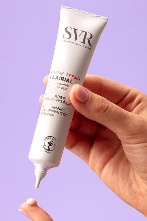 Mano che tiene un tubetto bianco di SVR Clairial Creme SPF50+ mentre eroga una goccia di crema su un dito.