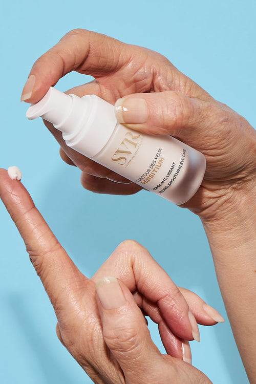 Mano che applica la crema contorno occhi SVR Densitium da un flacone bianco opaco su sfondo azzurro.