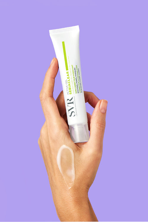 Hand holding a white SVR Sebiaclear Active Gel tube with a clear gel swatch on the skin against a purple background.