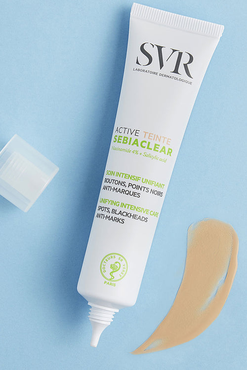 Tubo bianco di SVR Sebiaclear Active Teinte con tappo aperto e una goccia di crema colorata su sfondo azzurro.