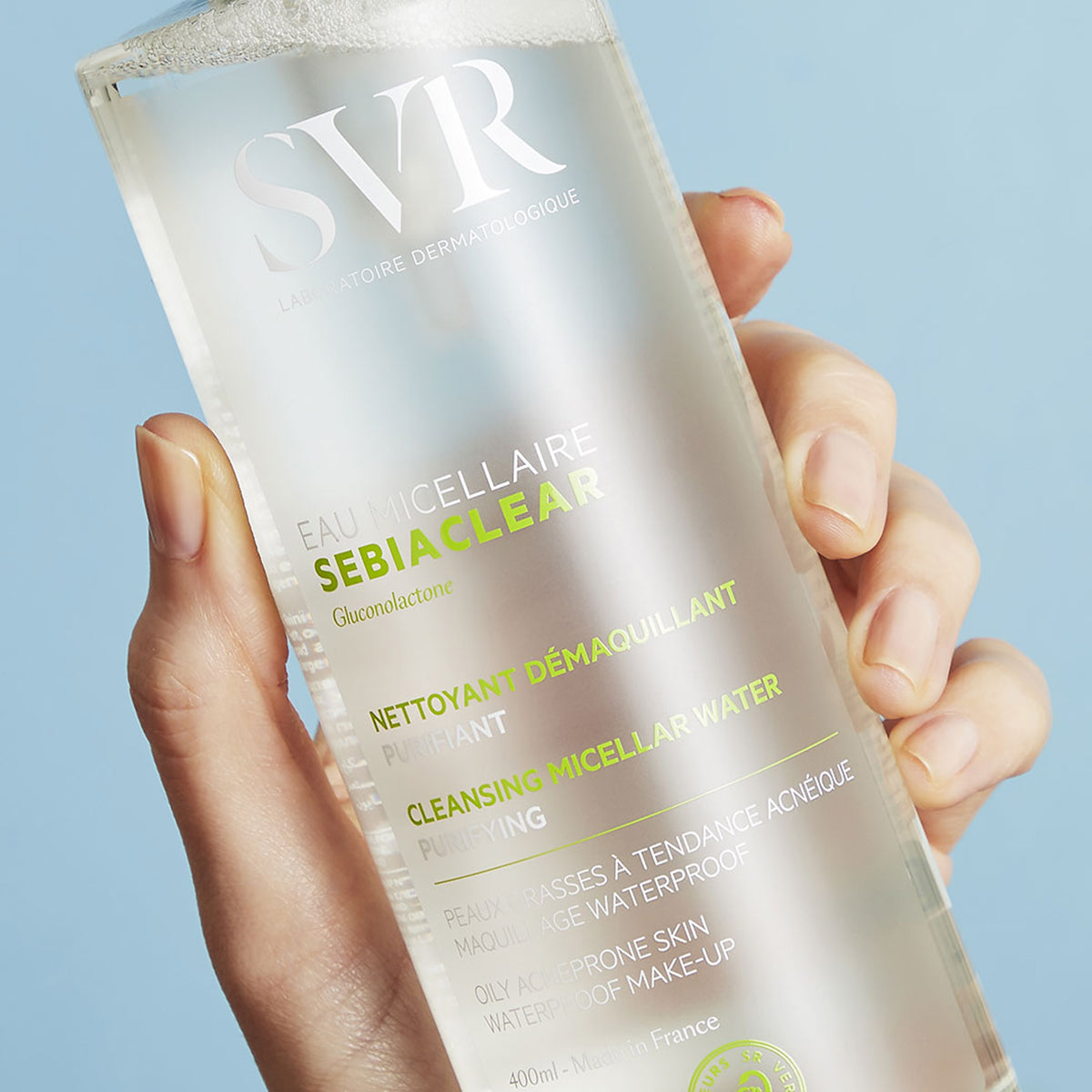 Flacone trasparente di SVR Sebiaclear Eau Micellaire da 400ml tenuto in mano su sfondo azzurro.