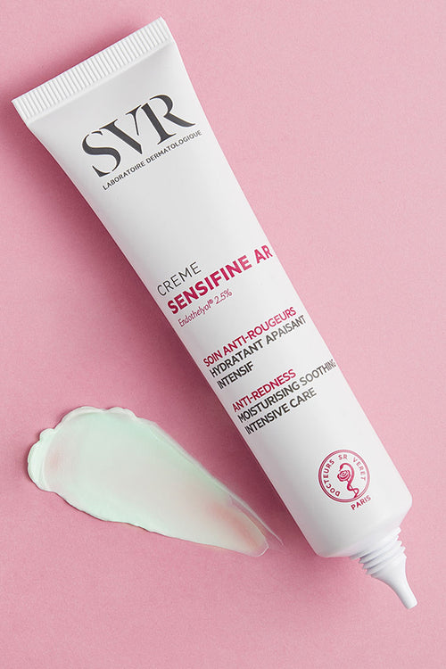 Tubo bianco di crema SVR Sensifine AR con una goccia di gel verde su sfondo rosa, ideale per pelli soggette ad arrossamenti.