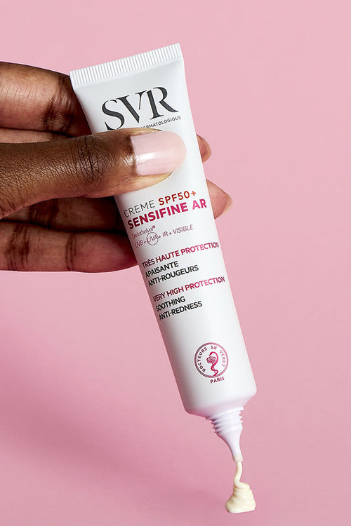 Tubo bianco di SVR Sensifine AR Crème SPF50+ tenuto in mano su sfondo rosa, con crema erogata dalla punta.