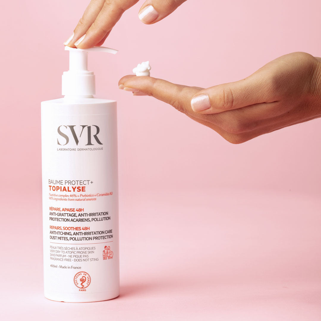 Flacone bianco con erogatore a pompa di SVR Topialyse Baume Protect+ mentre una mano preleva una dose di crema su sfondo rosa.