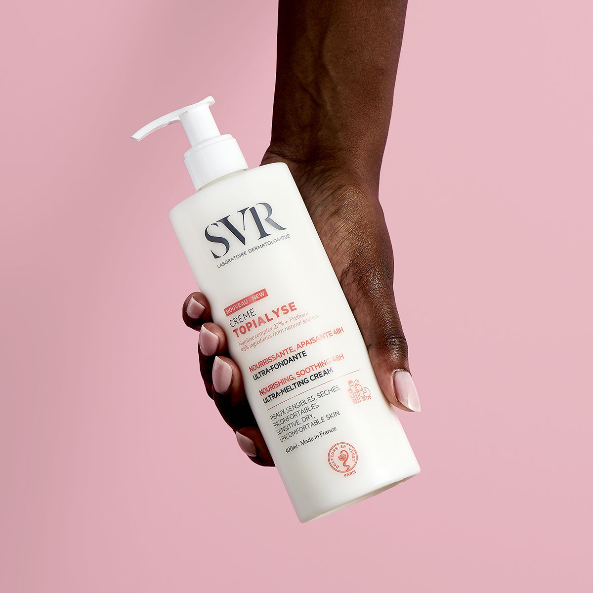 Mano che tiene un flacone bianco da 400ml di crema idratante SVR Topialyse con erogatore a pompa su sfondo rosa.