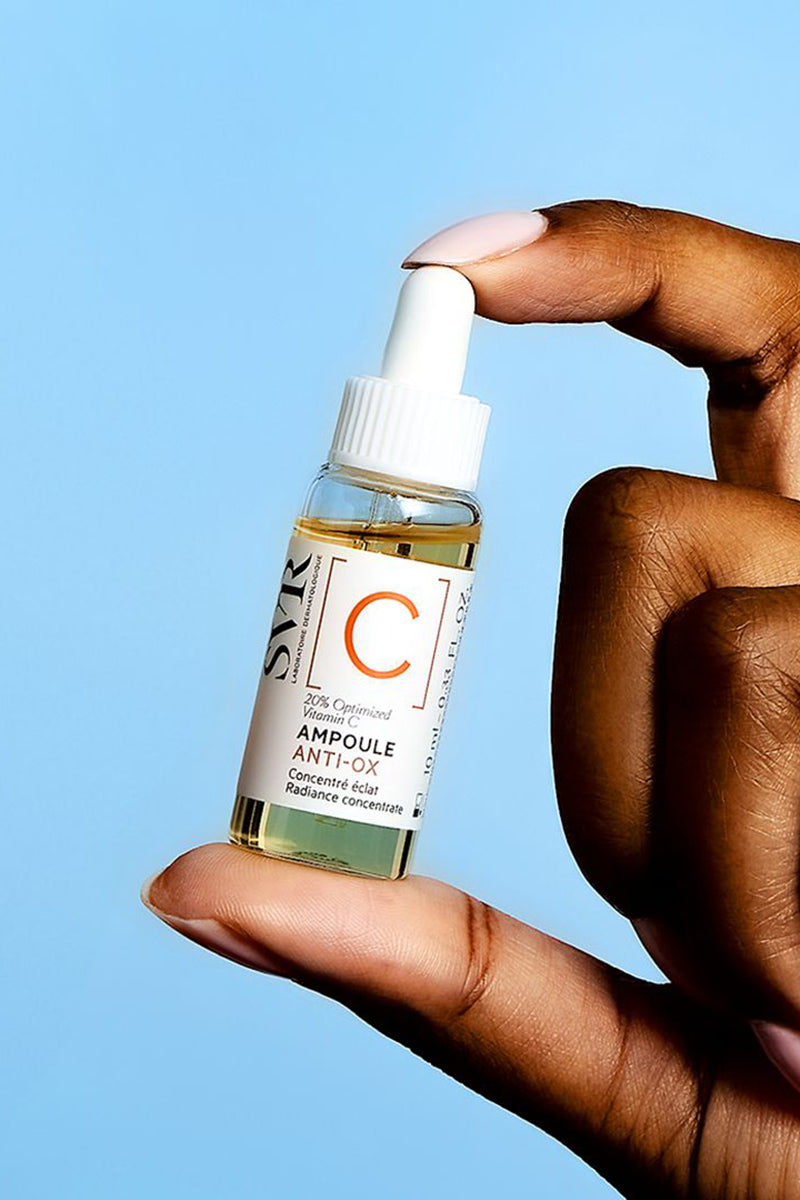 Mano che tiene un flacone di siero SVR Ampoule Anti-Ox con vitamina C su sfondo azzurro.