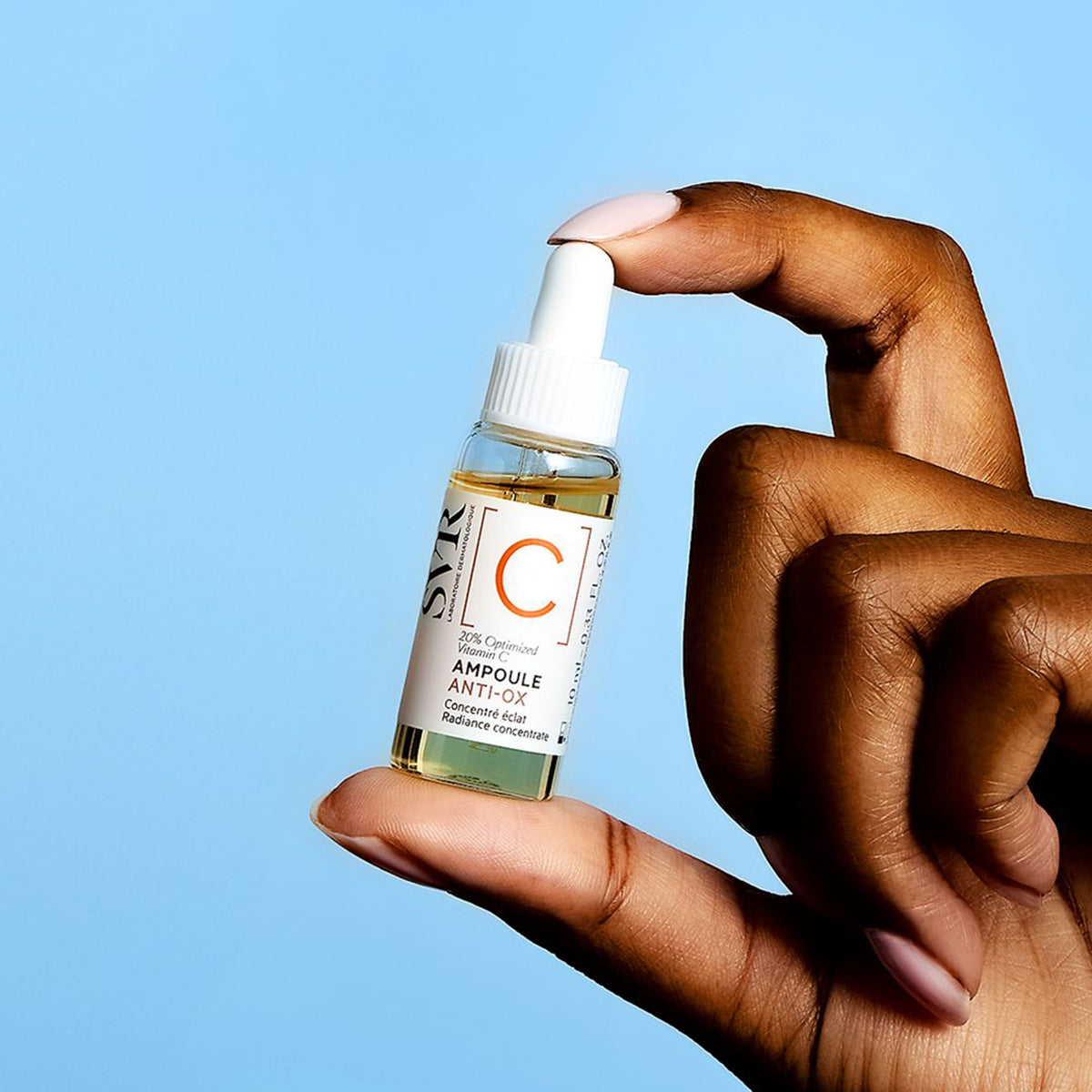 Mano che tiene un flacone di siero SVR Ampoule Anti-Ox con vitamina C su sfondo azzurro.