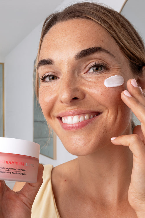 Woman smiling while applying SVR Ceramides Biotic regenerating nourishing balm from a pink glass jar to her cheek.
