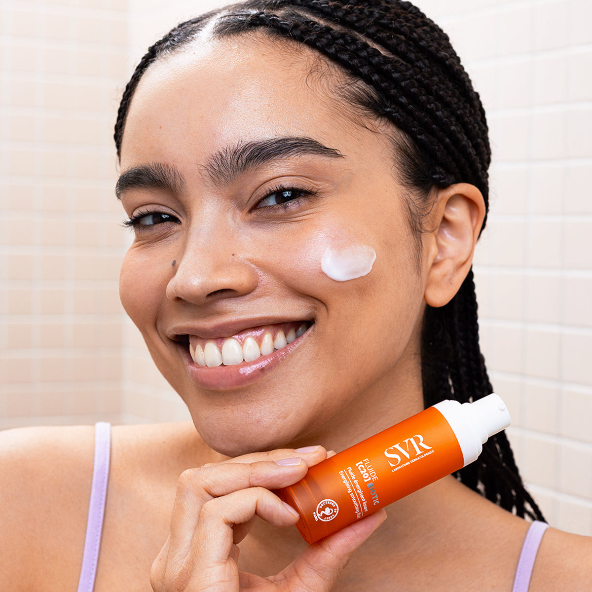 Smiling woman holding an orange SVR Fluide [C20] Biotic pump bottle with a dab of white cream on her cheek.