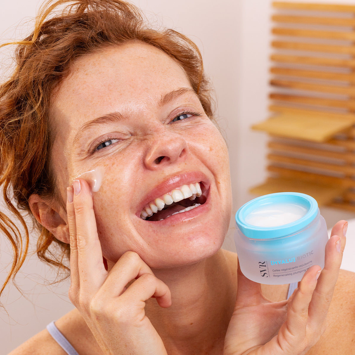 Donna sorridente applica la crema viso SVR [HYALU] Biotic dal vasetto in vetro satinato con tappo azzurro.