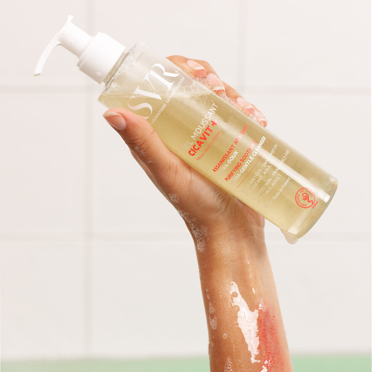 Flacone trasparente con dosatore di SVR Cicavit+ Gel Moussant tenuto in mano su sfondo piastrellato bianco.