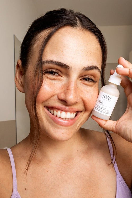 Donna sorridente tiene in mano il flacone di siero SVR Clairial Ampoule con contagocce in un bagno luminoso.