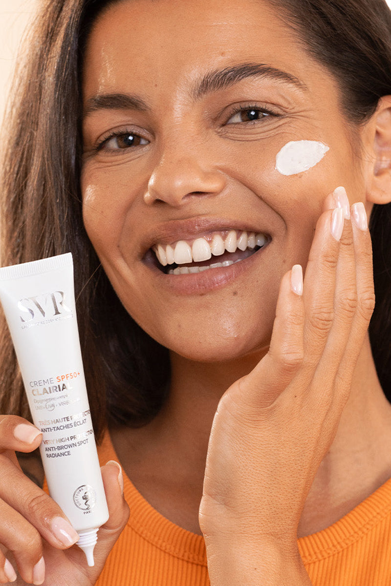 Donna sorridente applica la crema SVR Clairial SPF50+ bianca sul viso per trattare le macchie cutanee.