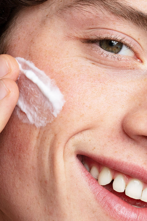 Primo piano di un giovane che applica una crema idratante bianca e leggera sulla guancia, sorridendo con naturalezza.