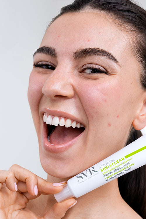 Smiling woman holding a white SVR Sebiaclear Active Gel tube with green accents in a bright bathroom setting.