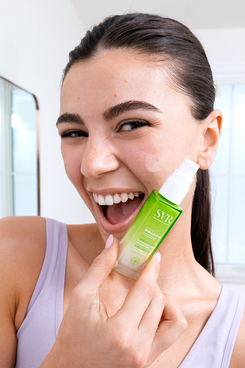 Donna sorridente tiene in mano il flacone verde di SVR Sebiaclear Sérum anti-imperfezioni in un bagno luminoso.