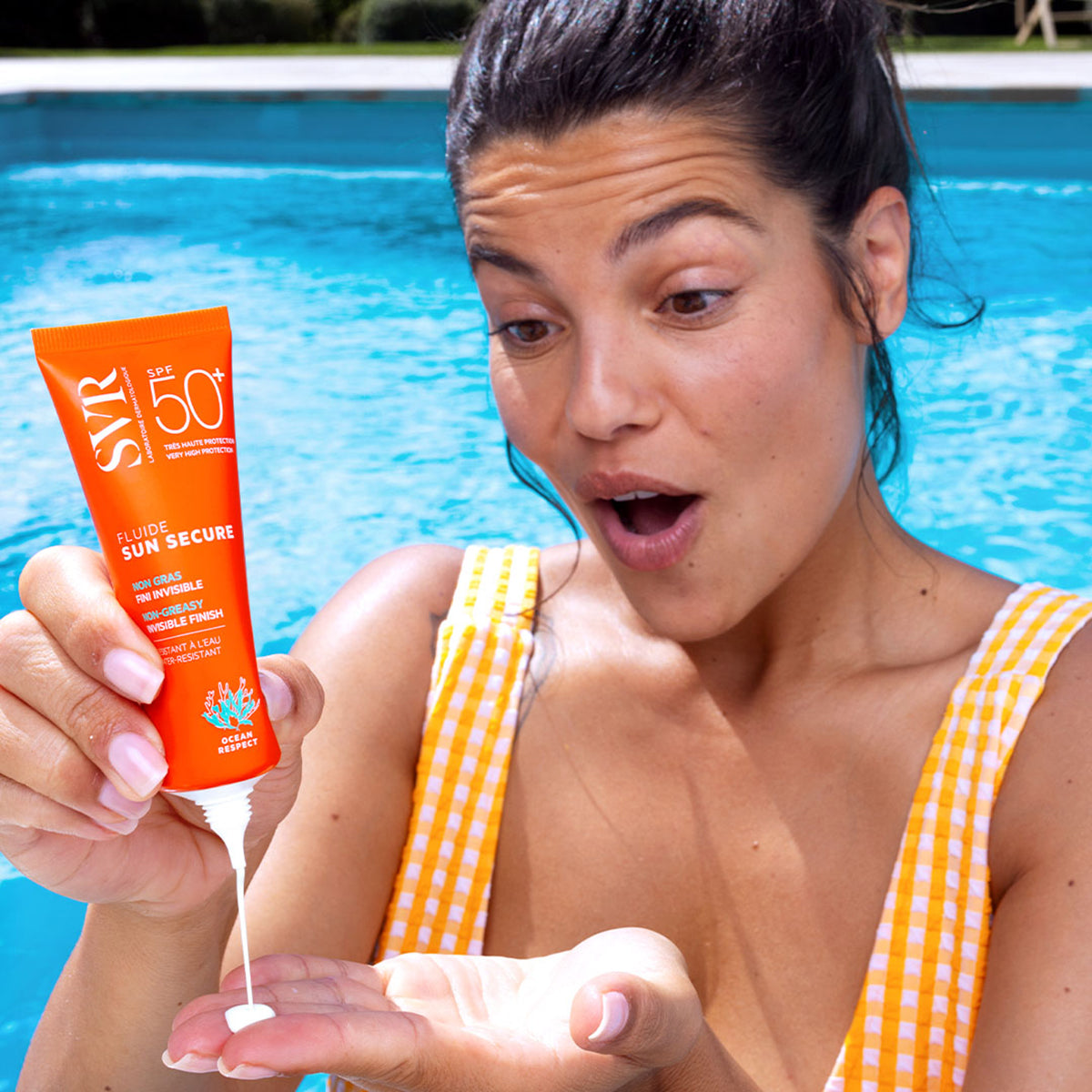 Donna in costume a quadretti applica la crema solare SVR Sun Secure Fluide SPF50+ a bordo piscina.