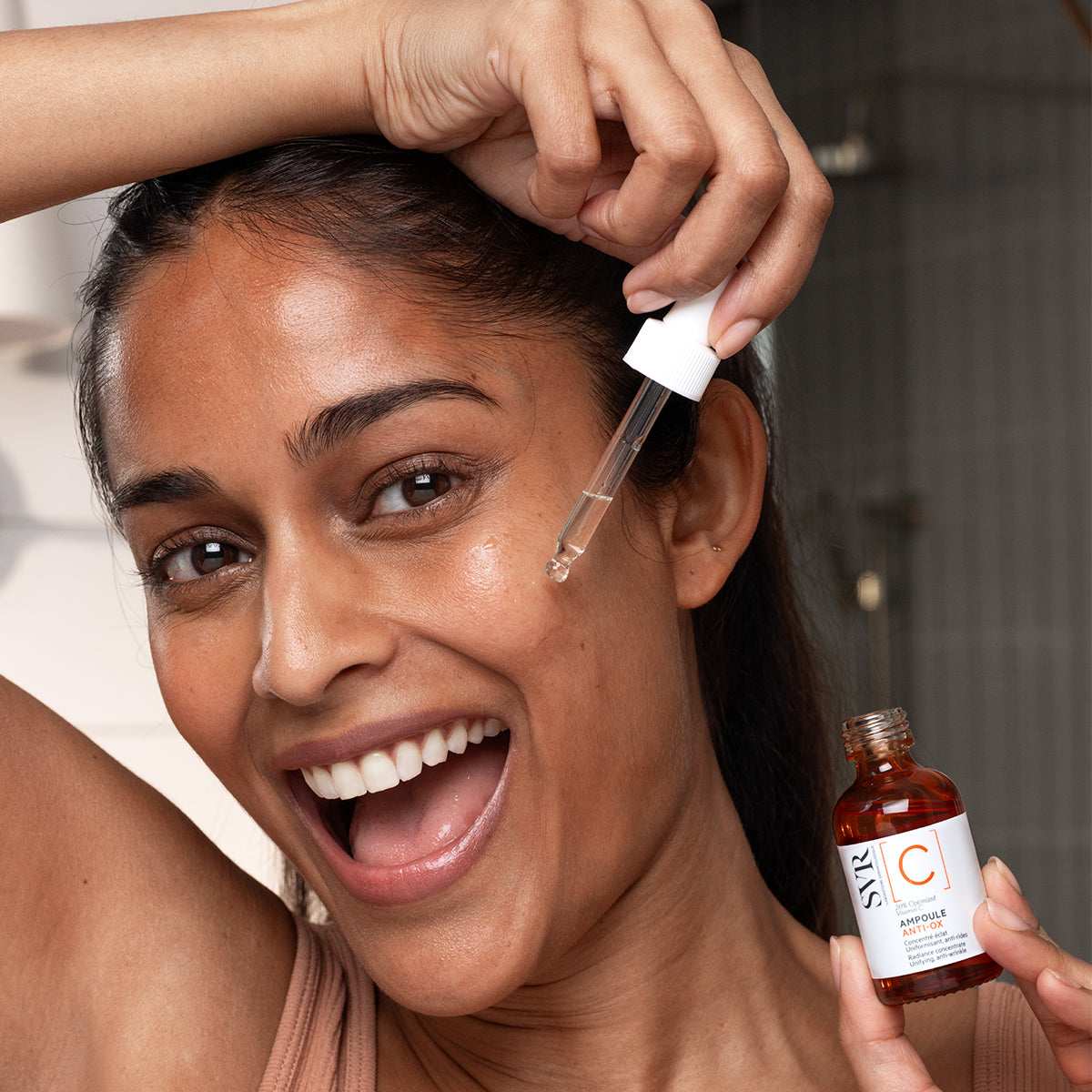 Donna sorridente applica il siero SVR Ampoule Anti-Ox con contagocce da una boccetta in vetro ambrato per una pelle luminosa.