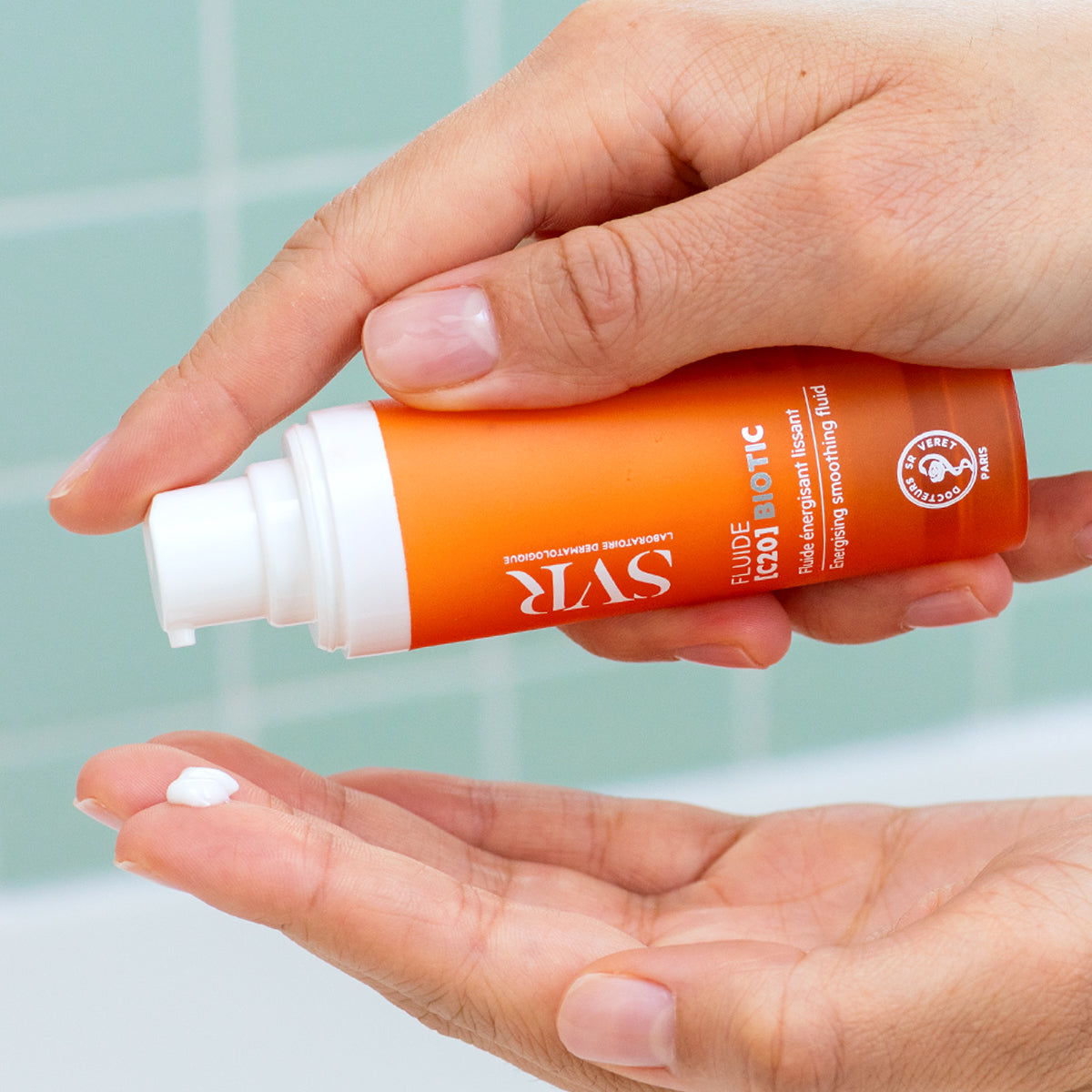 Hand holding an orange SVR Fluide [C20] Biotic pump bottle, dispensing a drop of white smoothing cream into the palm.