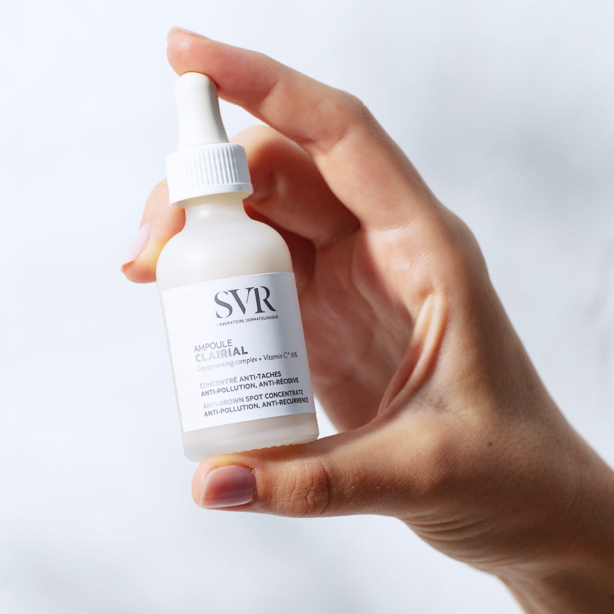 Mano che tiene un flacone di siero SVR Clairial Ampoule in vetro satinato con contagocce bianco su sfondo chiaro.